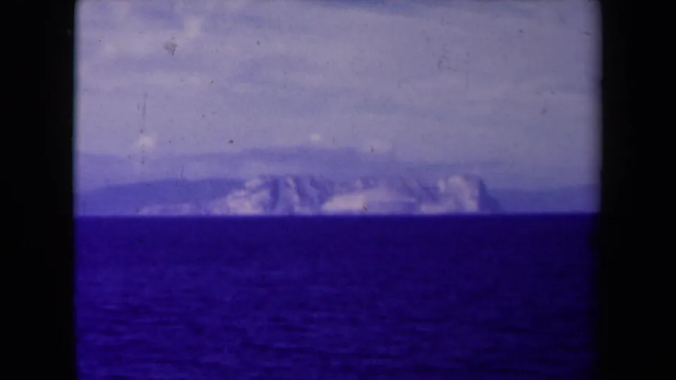 A final 8mm frame holds on rugged distant mountains across the sea, with cloudy sky, film grain, and dark vertical frame edges.