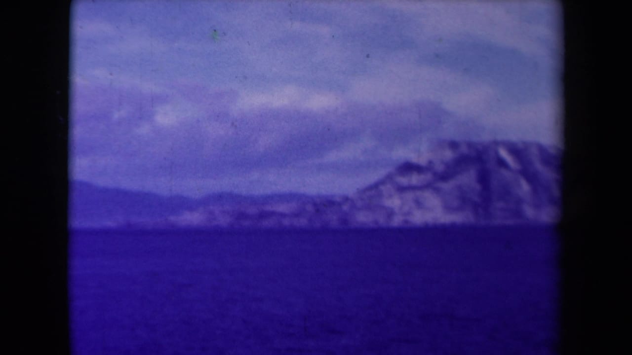 Remote cliff shoreline across blue water with mountains or ridges in the distance.