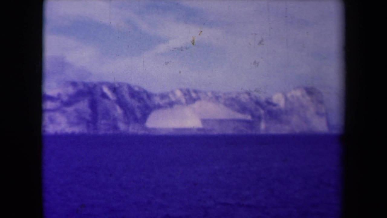 Purple-shifted 8mm frame from 1945 France showing dark water below a rugged icy shoreline and pale sky.