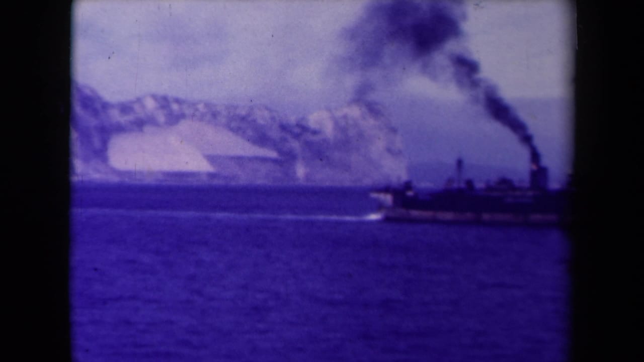 A distant smoking steamship moves across cold blue water near a glacier-like shoreline in 1945 France home-movie footage.