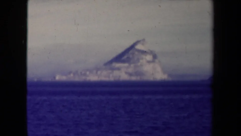 1945 8mm frame with a distant rocky coastal mass on the horizon, seen across dark blue water with soft archival color.