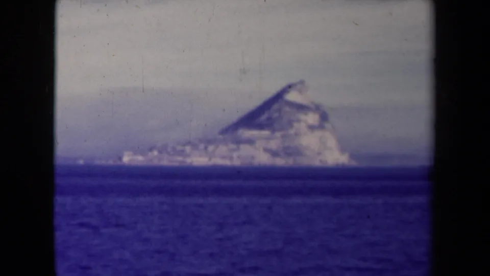 1945 France 8mm frame showing a rugged mountain-like coastal landform beyond open water, with visible grain and soft focus.