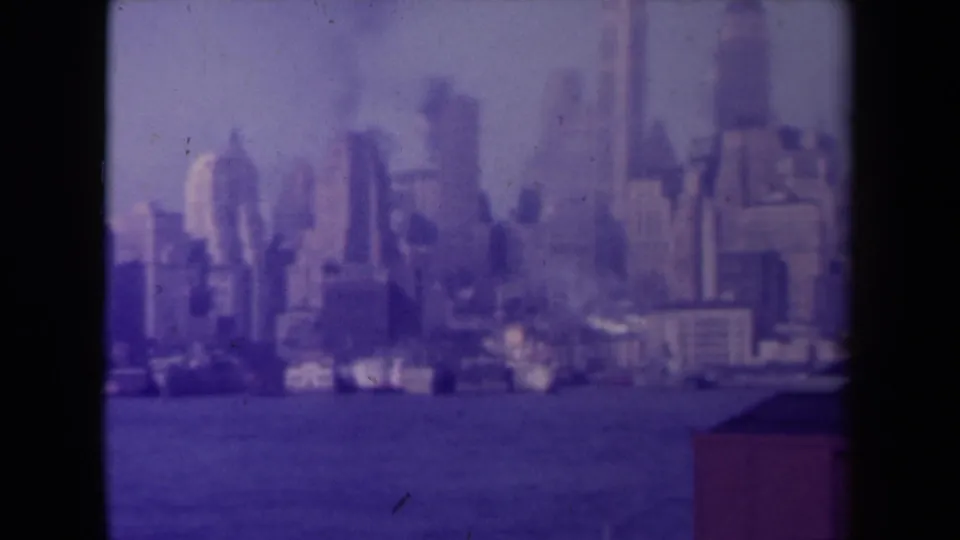 1940s home-movie frame of a soft, purple-tinted city skyline seen across water with dark vertical borders.