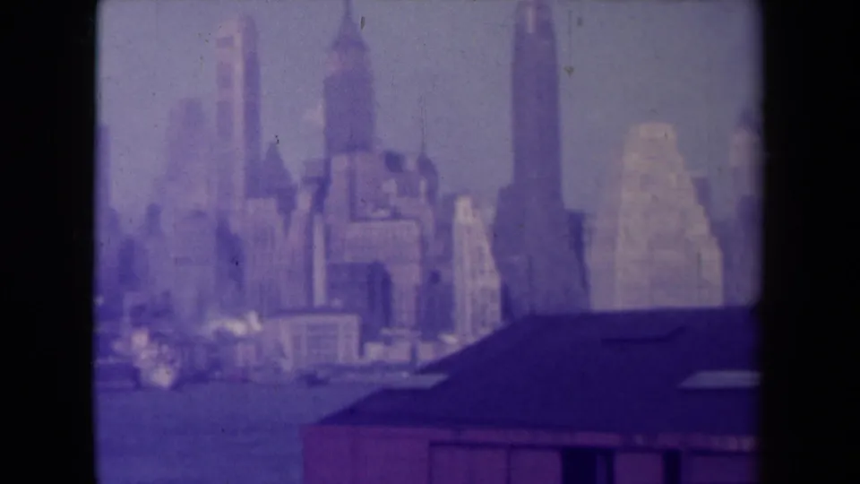 Archival 8mm frame showing a distant city skyline across water, partly blocked by a dark foreground roof.