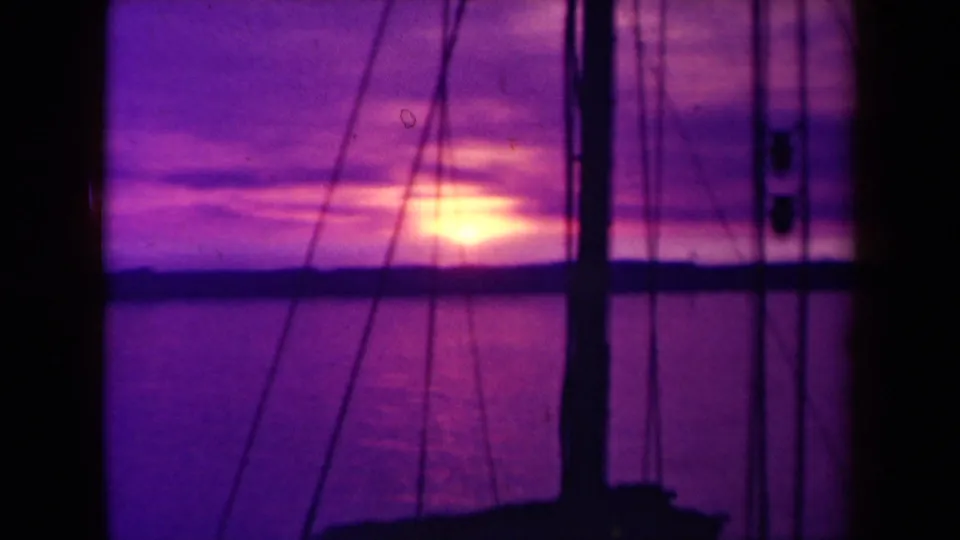 1940s France 8mm frame showing a warm sun near the sea horizon, seen past boat rigging and dark vertical mast shapes.