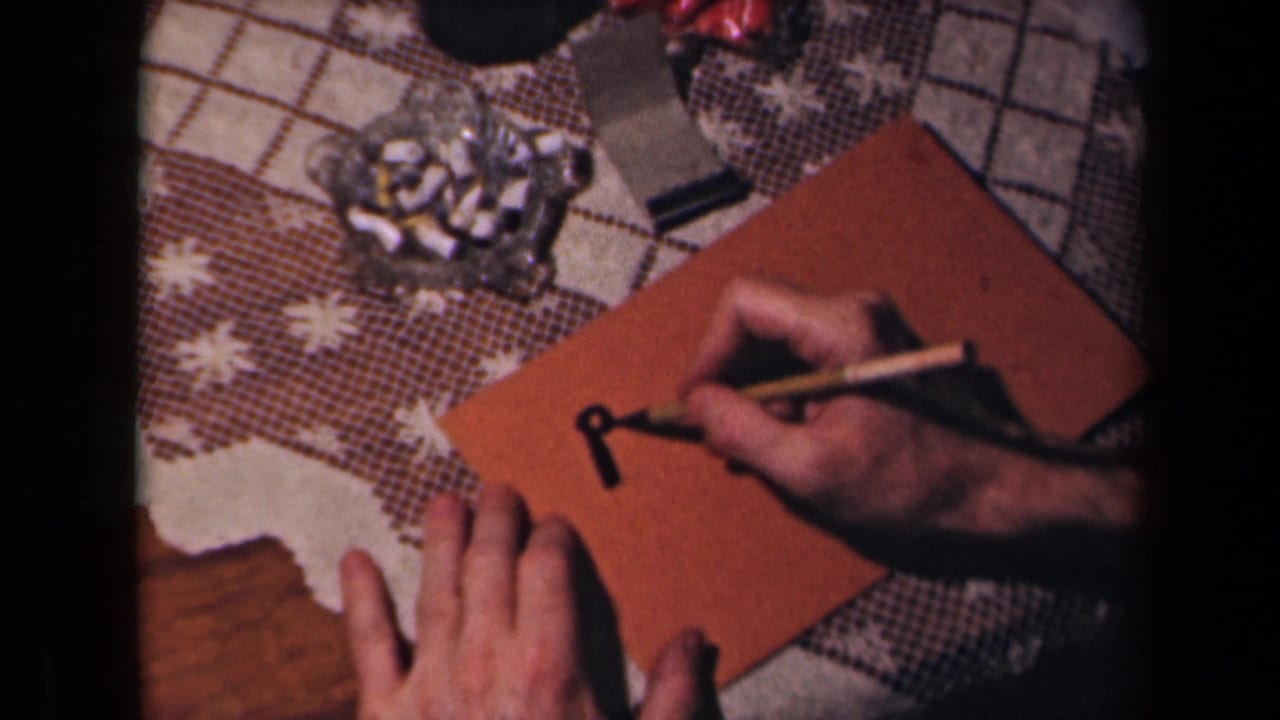 Hands begin writing on a brown envelope beside a glass ashtray on a patterned tablecloth.