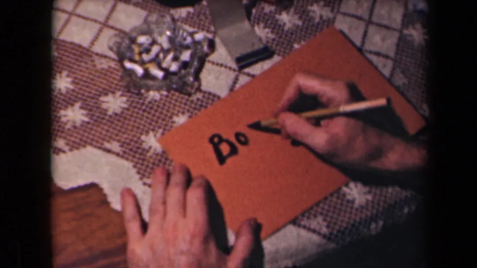 A close 1945 French home-movie frame shows a hand writing Bo on a brown envelope beside an ashtray on a patterned table.
