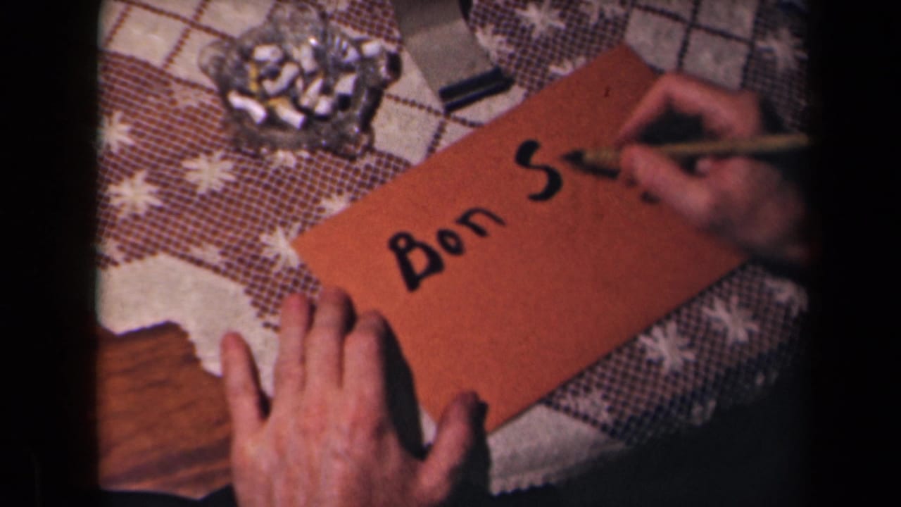Hands hold a brown card while a dip pen writes the start of “Bon Soir” on a patterned table.