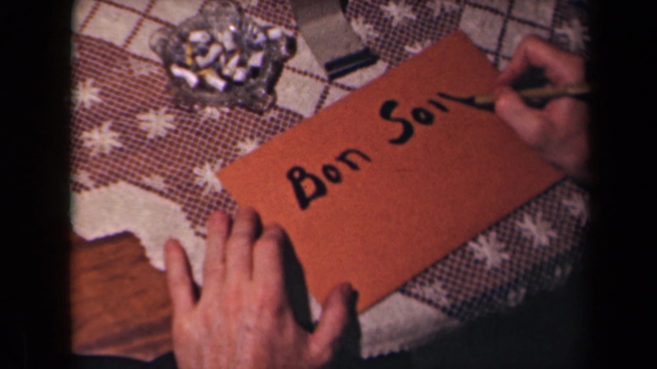 A hand continues writing “Bon Soir” on a brown card near a glass ashtray and tablecloth.