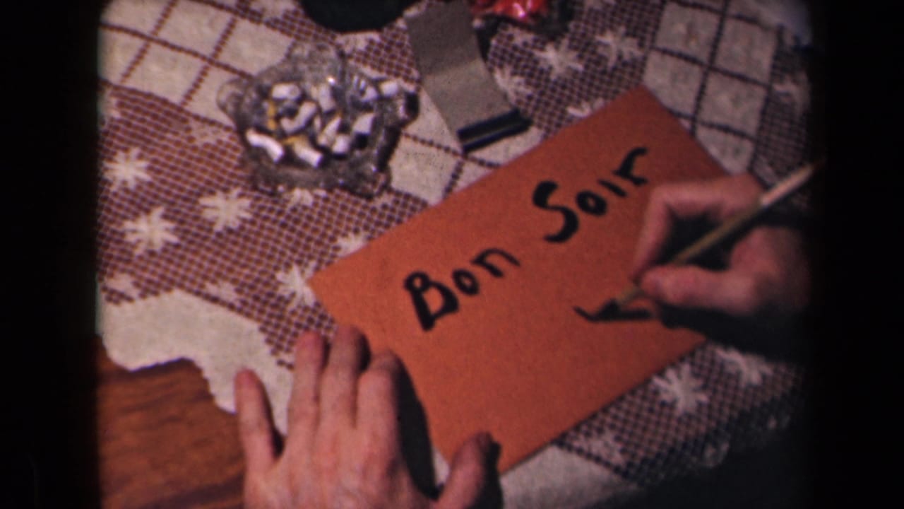 Hands write Bon Soir on an orange envelope beside a glass ashtray on a patterned tablecloth.