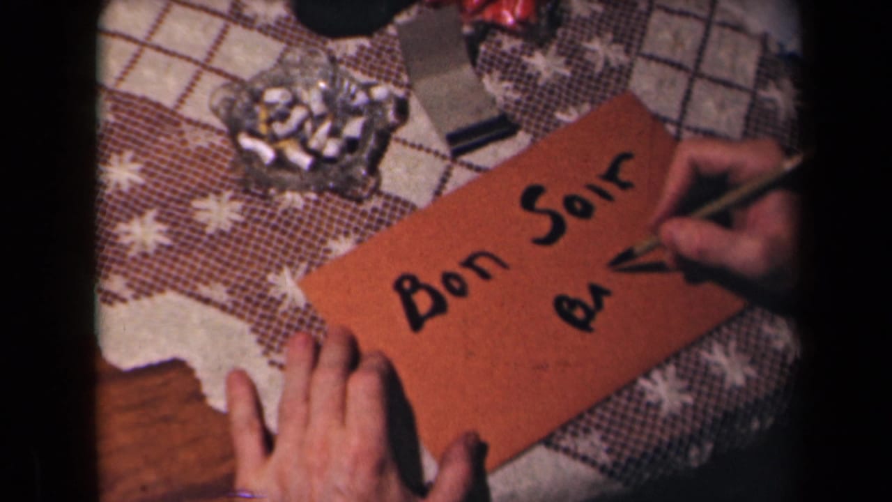 A close 8mm view shows Bon Soir written on an orange envelope while a hand adds more black script on a domestic tabletop.