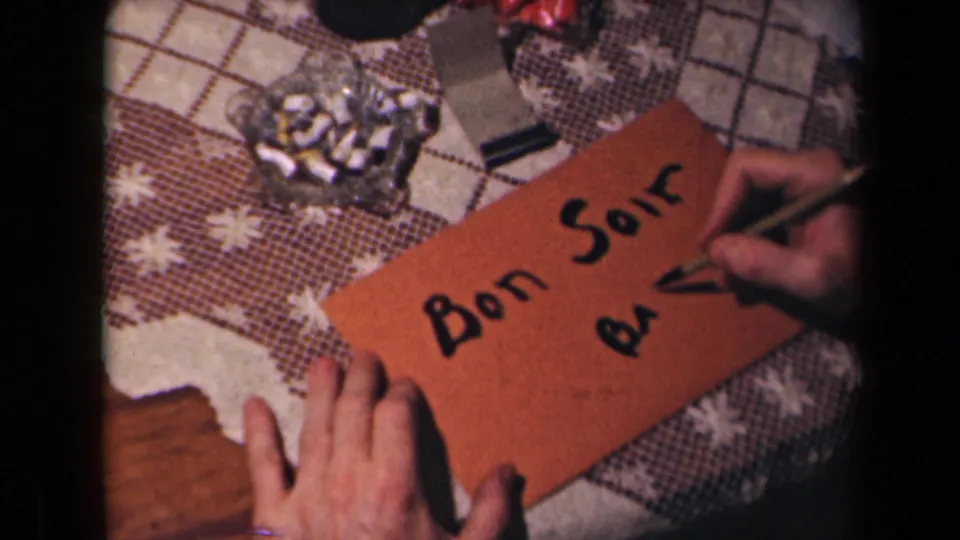 A close 8mm view shows Bon Soir written on an orange envelope while a hand adds more black script on a domestic tabletop.