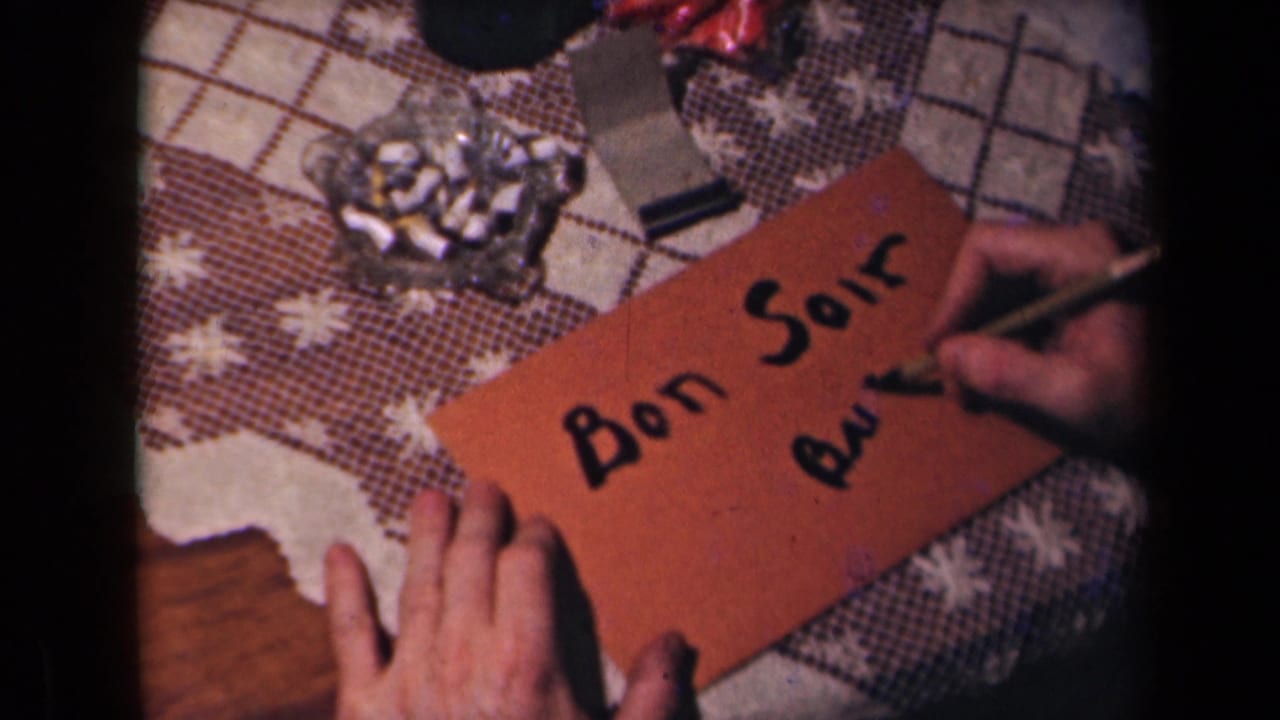 The handwritten Bon Soir remains visible as a hand adds lower text on the orange envelope, with ashtray and tablecloth in view.