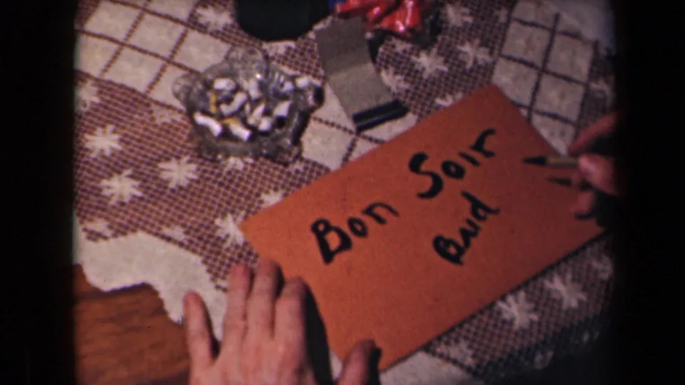 In 1945 France home-movie footage, hands steady a brown card as a personal note is written beside an ashtray on a patterned tablecloth.