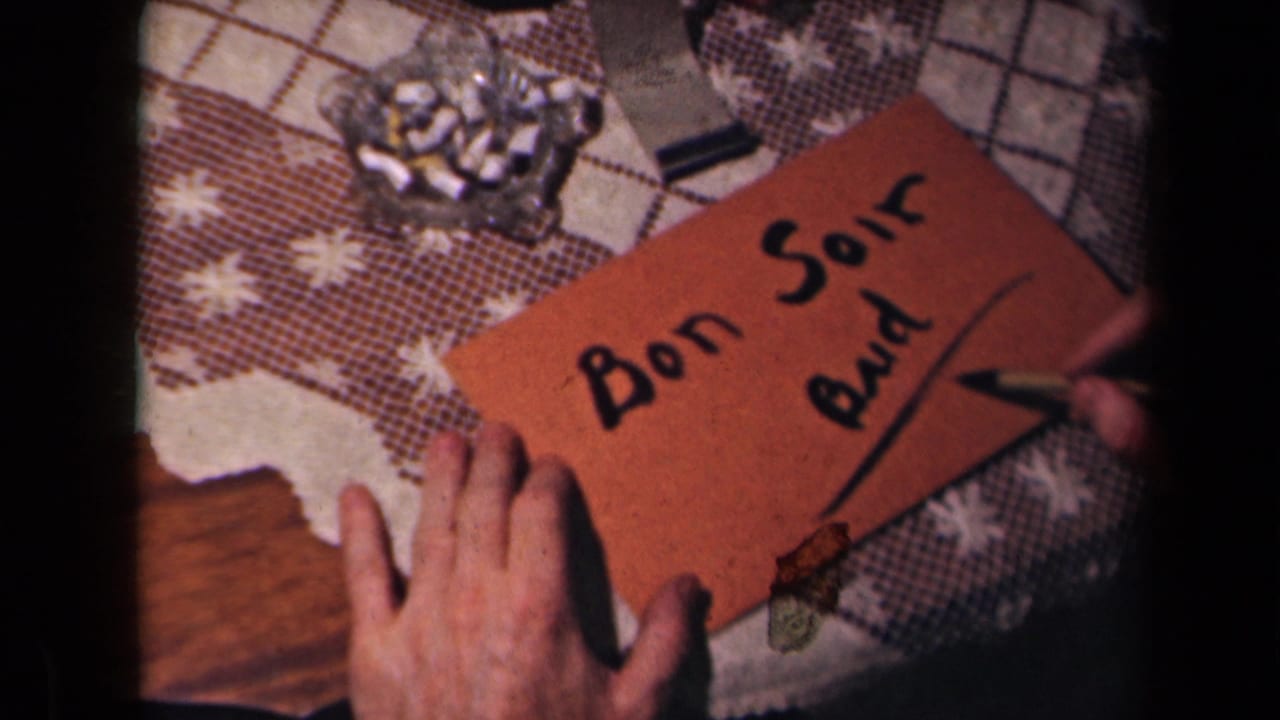 The brown card with "Bon Soir Bud" fills the frame as a hand makes another mark near the lower edge on a patterned tabletop.