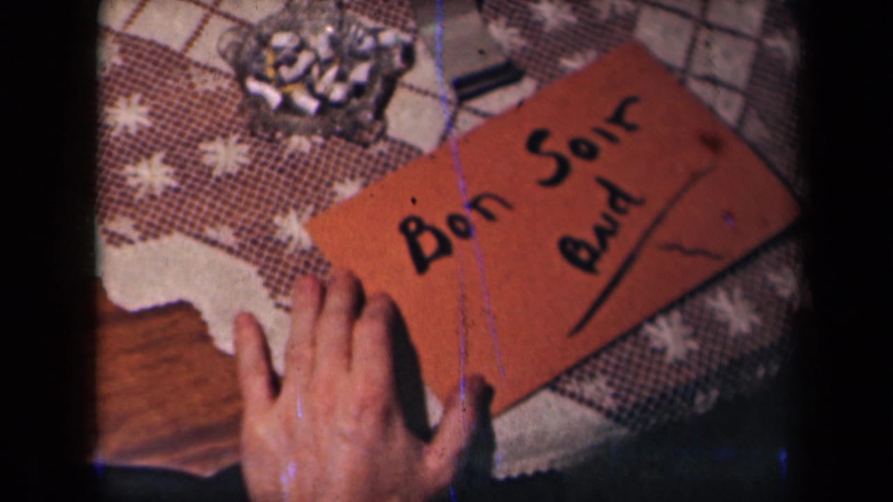 The completed handwritten card reading "Bon Soir Bud" rests on a patterned tablecloth, with hands and a glass ashtray visible nearby.