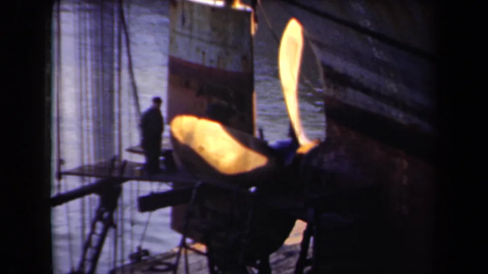 A large illuminated propeller dominates the dockside view, with a worker silhouette, hull plating, rigging, and water nearby.