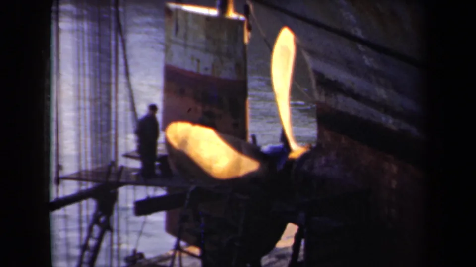 1946 Netherlands 8mm frame showing a worker silhouetted near a huge sunlit ship propeller beside a large vessel hull.