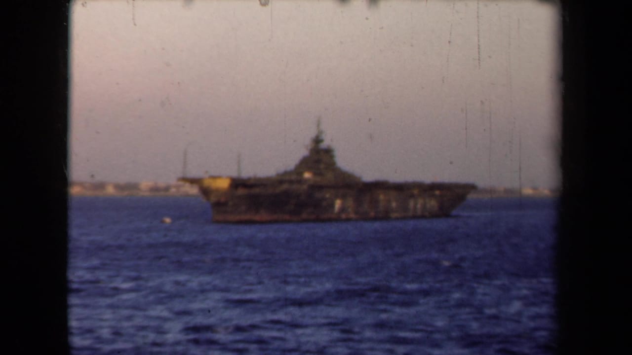Large dark naval ship on blue water with a distant shoreline and pale sky.