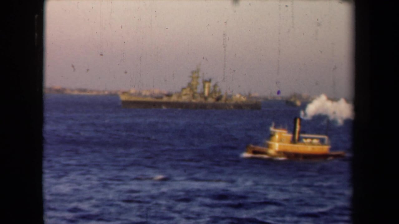 Small yellow tugboat on blue water with a distant gray warship behind it.