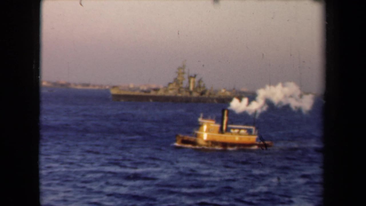 Yellow tugboat with white smoke crossing water in front of a distant warship.