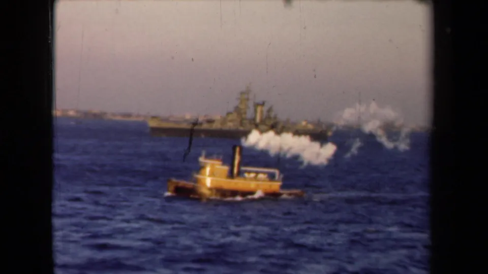 The small yellow tugboat continues across choppy water beneath white smoke, with a distant naval vessel still visible near the horizon.