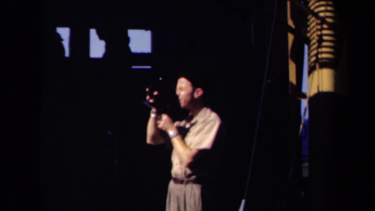 In 1946 Netherlands home-movie footage, a man in a pale shirt and dark hat appears to film with a small camera inside a shadowed interior with bright high windows.