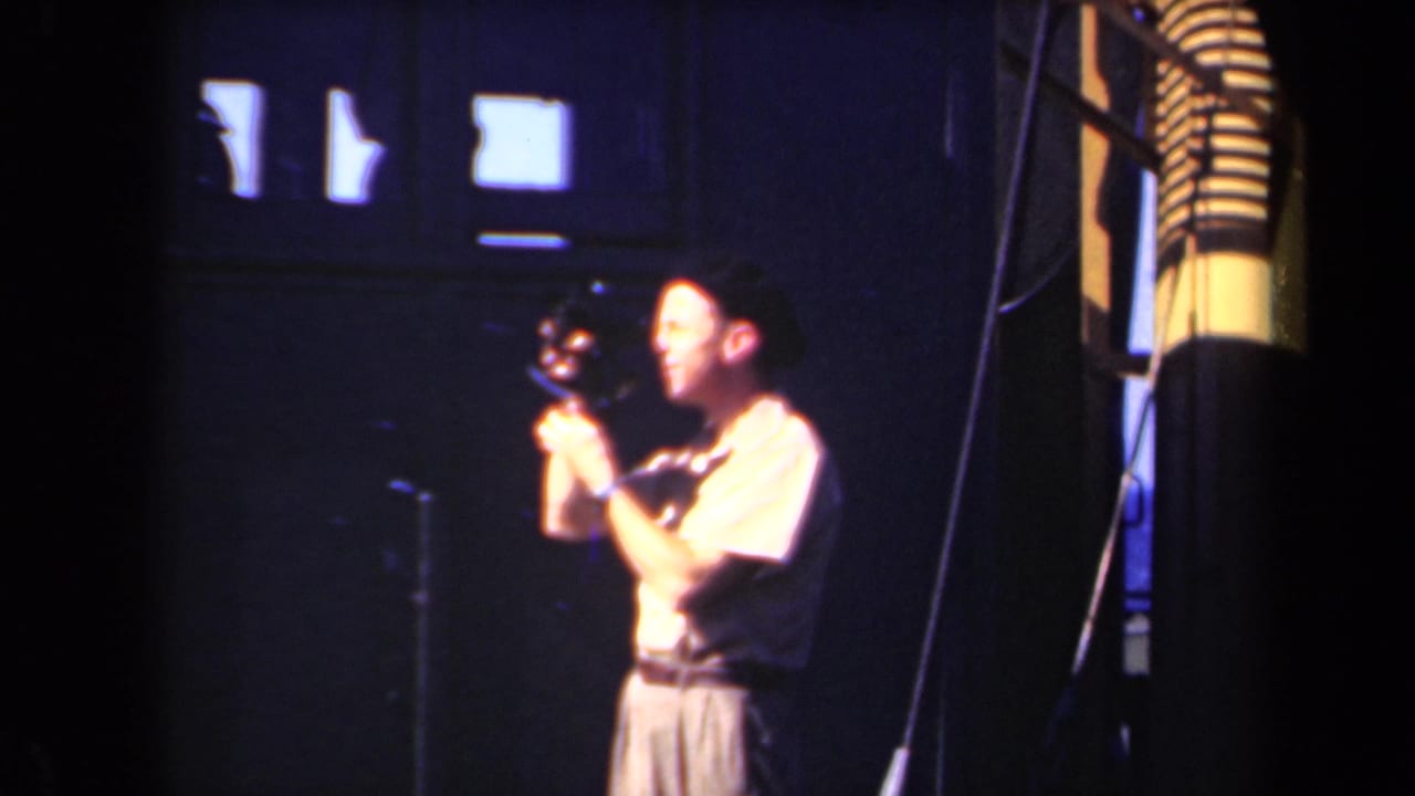 A man in profile, identified by metadata as filmed in the Netherlands in 1946, holds a small camera near his eye against a dark interior background with lit rectangular windows.