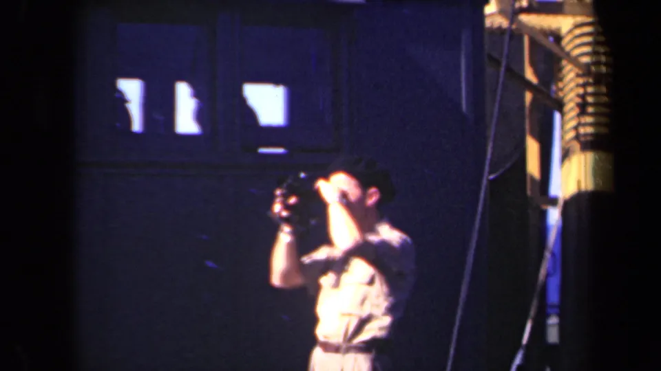 A man in pale clothing holds a small camera up to his eye in 1946 Netherlands home-movie footage, framed by dark interior walls, high windows, and a striped column at the right edge.
