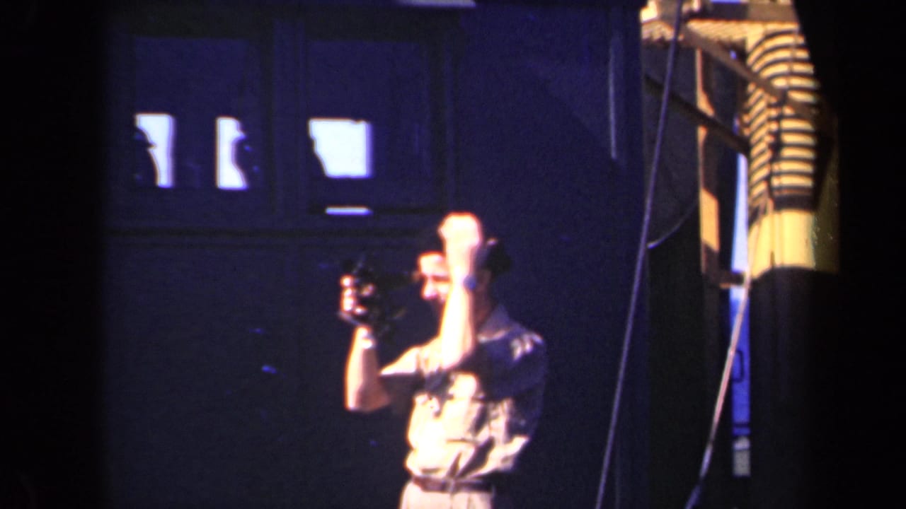 Man in a light shirt and dark cap raises a small movie camera inside a dim industrial space.
