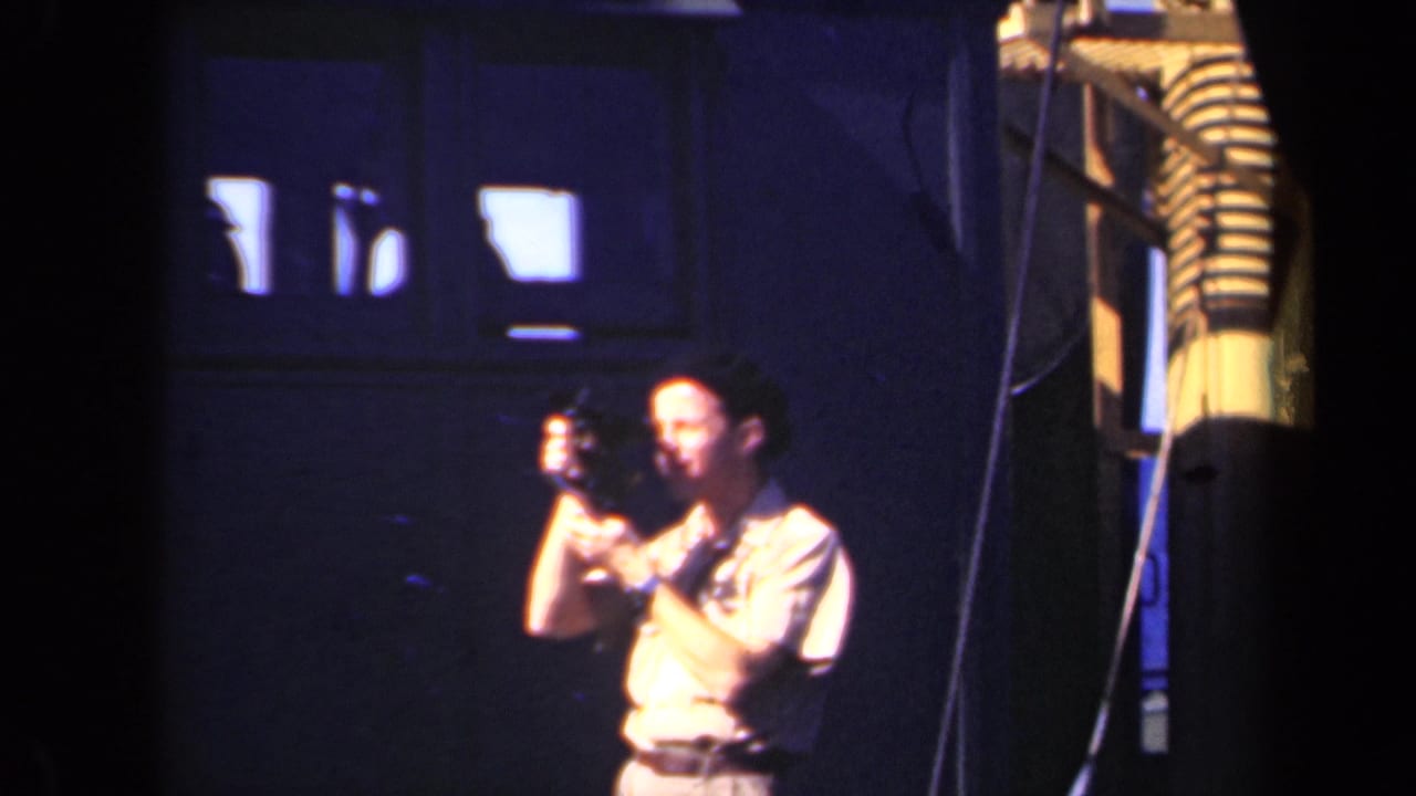 A man in a cap films inside a dim industrial interior, with bright windows and machinery or structural elements nearby.