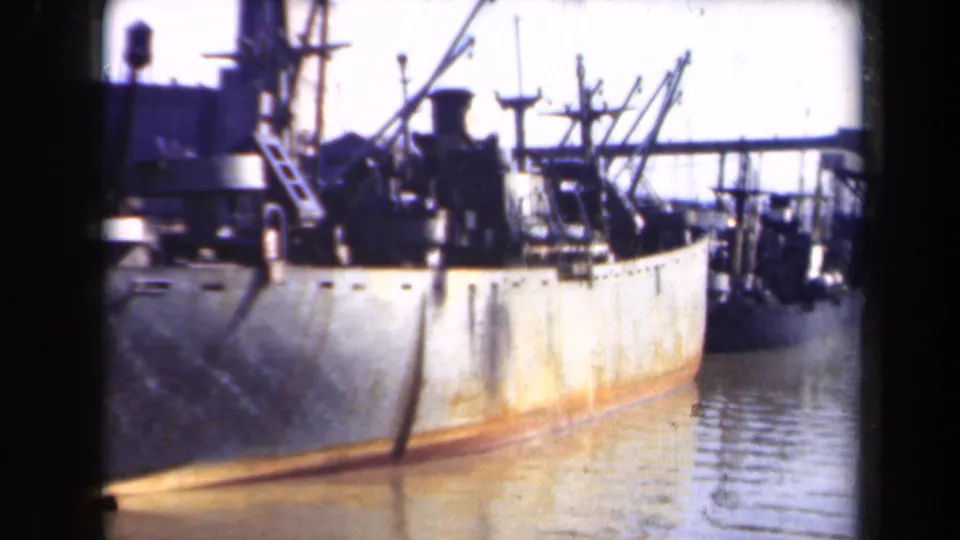 A large weathered commercial vessel appears docked in calm channel water, with rigging, deck equipment, and a bright harbor background.