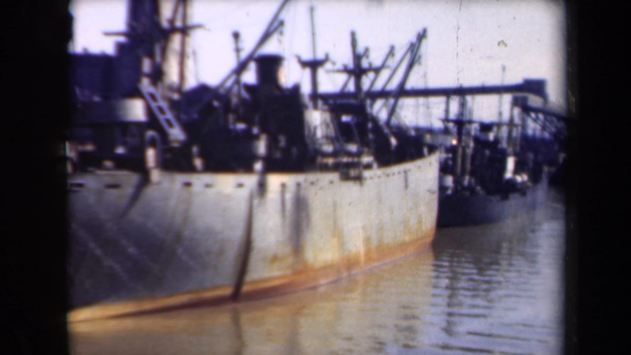 Rust-stained cargo ship moored in calm harbor water beside dock equipment.