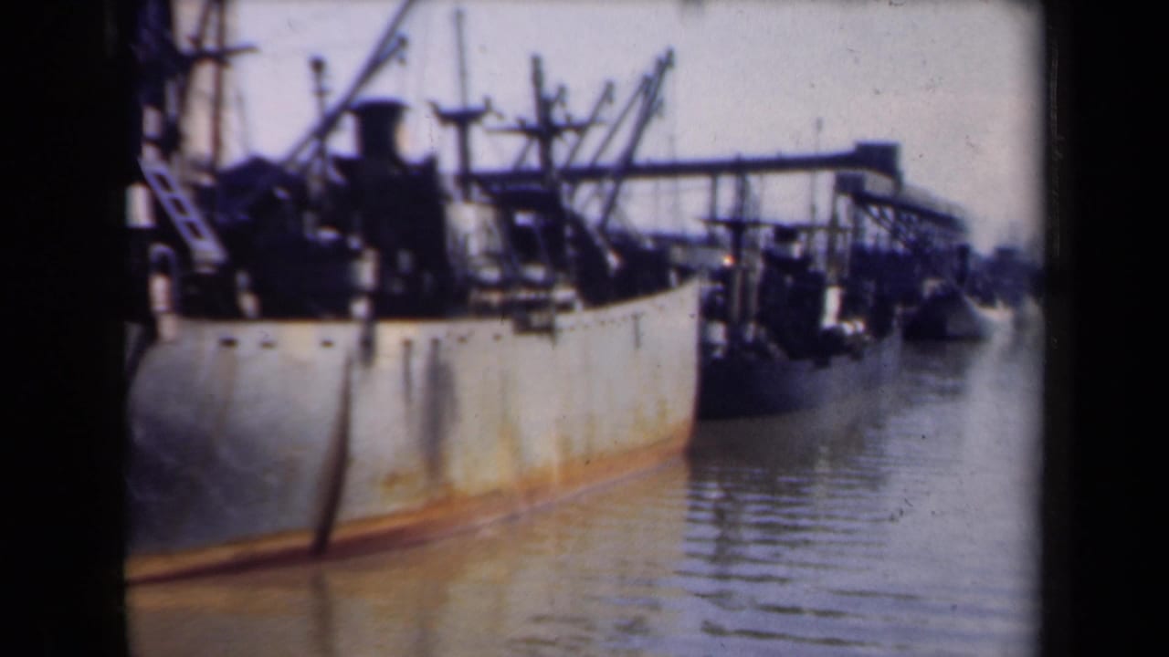 1946 Netherlands 8mm frame of a moored cargo vessel with cranes, railings, and rigging above a rust-stained hull in still harbor water.
