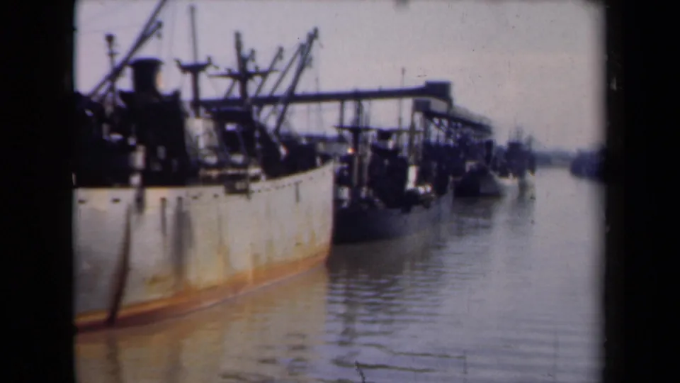 1946 Netherlands 8mm frame showing the side of a rusted cargo ship at dock, with calm water and another vessel or harbor form in the distance.