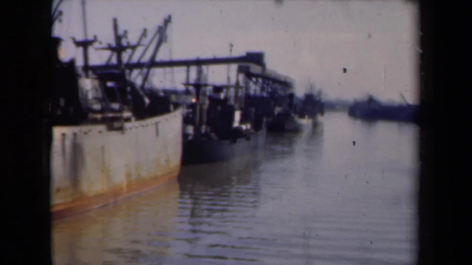 1946 Netherlands 8mm frame of a rusted cargo ship moored along a quiet dock, with deck equipment, calm water, and distant harbor shapes beyond.