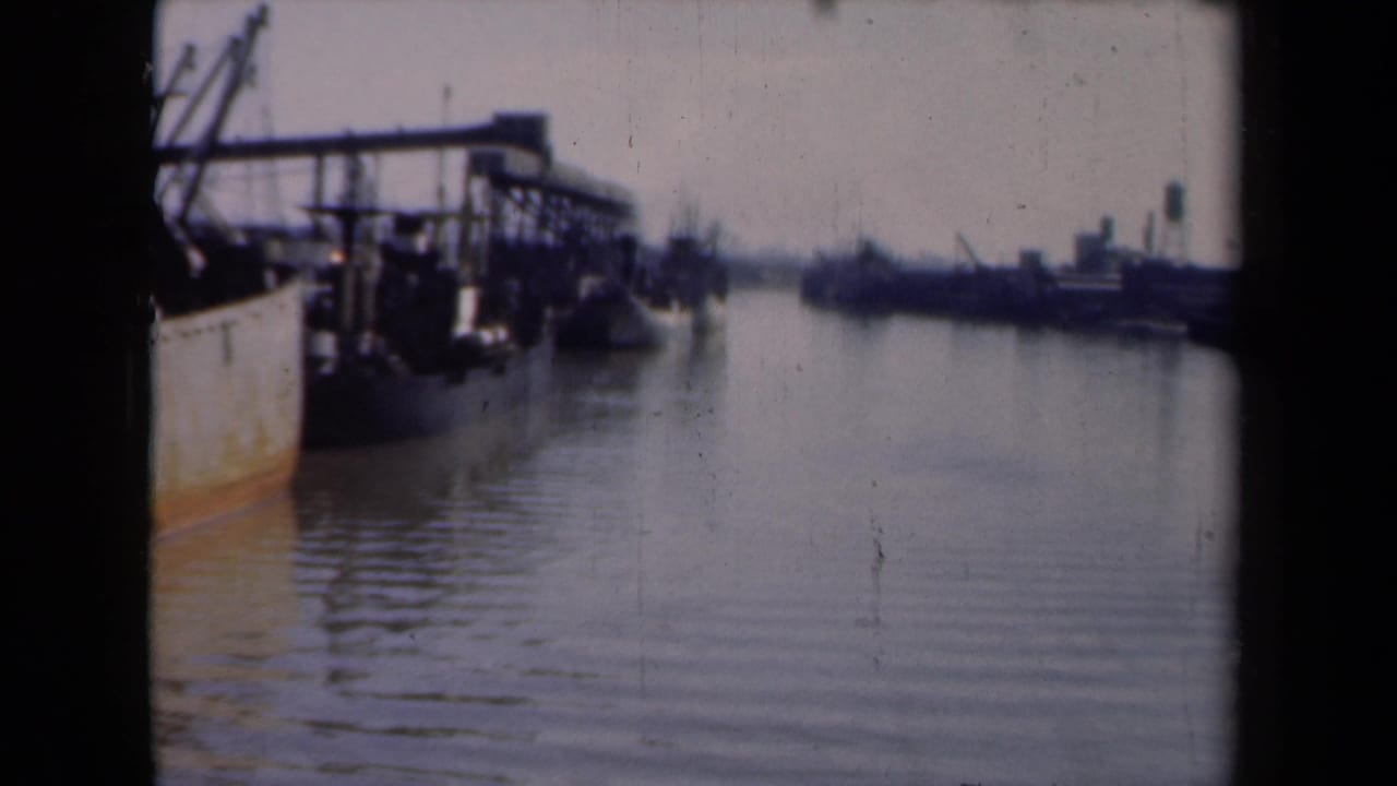 Dutch harbor scene from 1946 showing dockside structures, moored vessels, calm water, and distant industrial ship silhouettes.