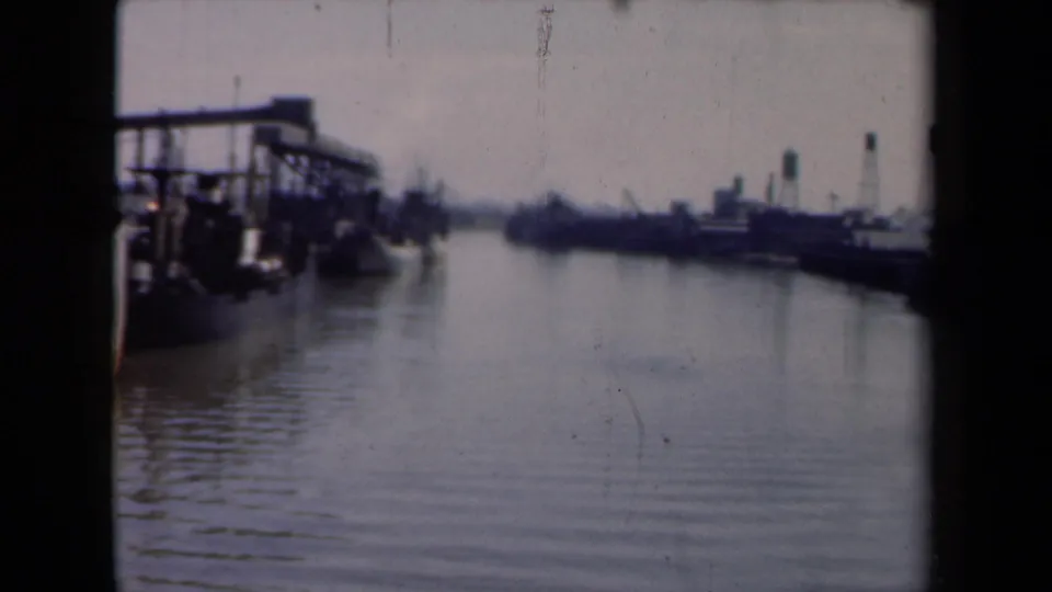 1946 Netherlands waterway view with dock structures, low moored ships, calm rippled water, and a distant industrial shoreline.
