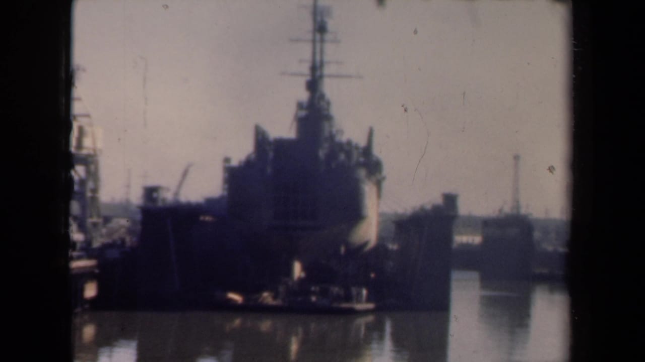 Large dark ship entering a harbor with cranes and dock structures nearby.