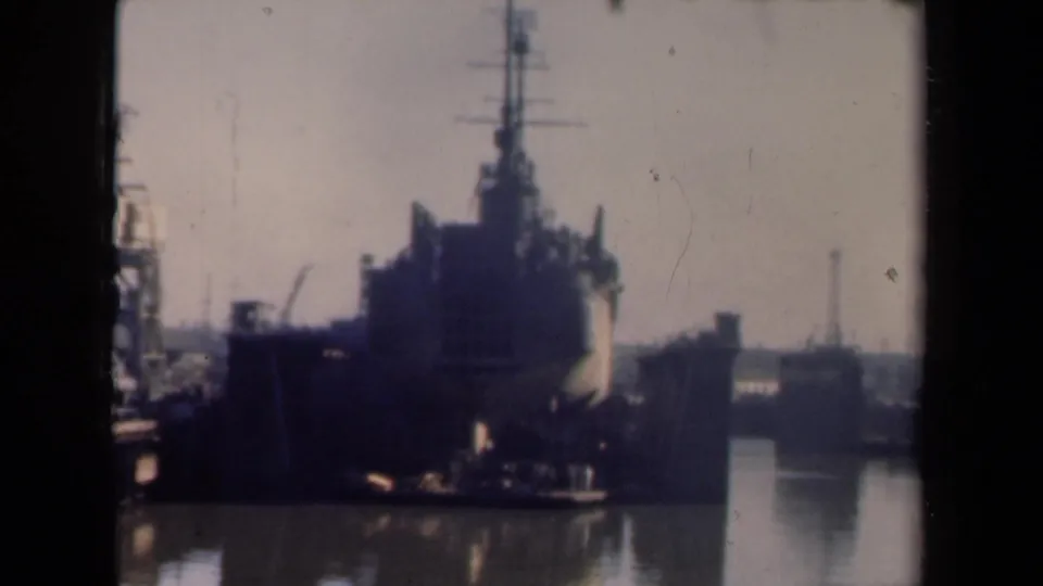 1946 Netherlands 8mm frame showing a large dark vessel approaching port, with cranes, calm water, and hazy shoreline structures.