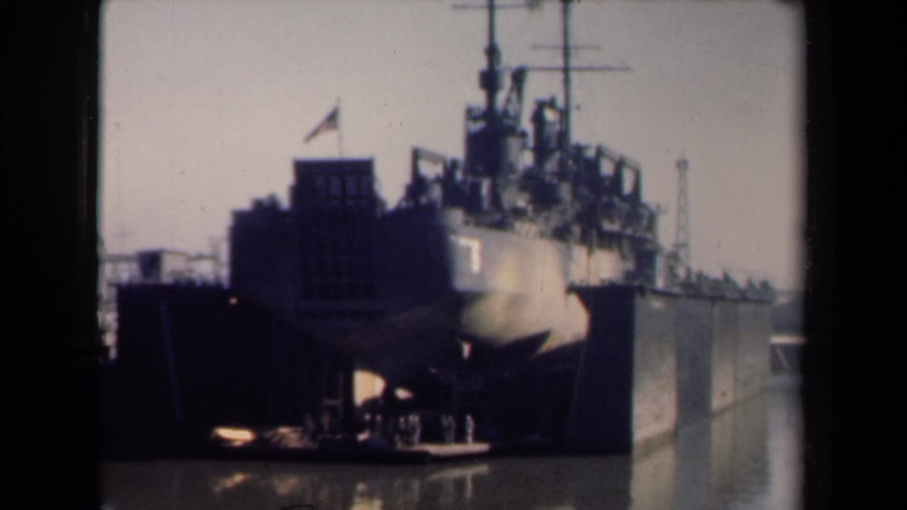 Large American military vessel in a Netherlands harbor, filmed from shore with dock structures and water in view.