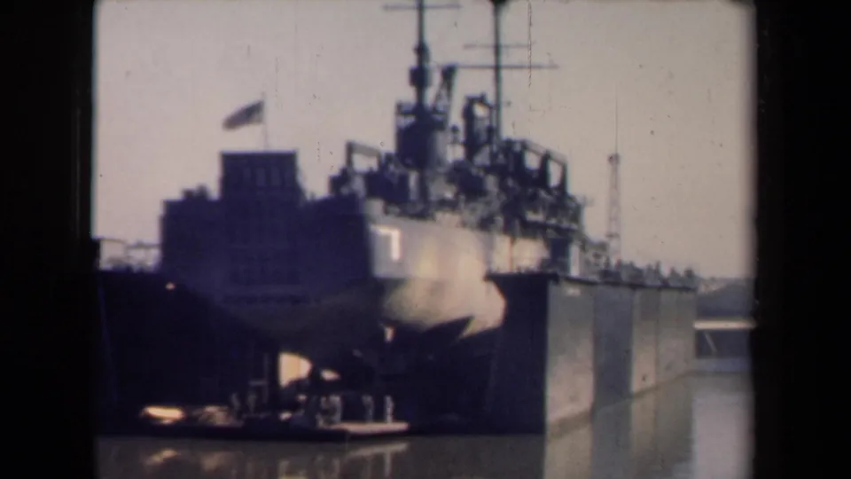 Soft 8mm frame of a docked American warship in a Netherlands harbor, with small people near the lower pier area.