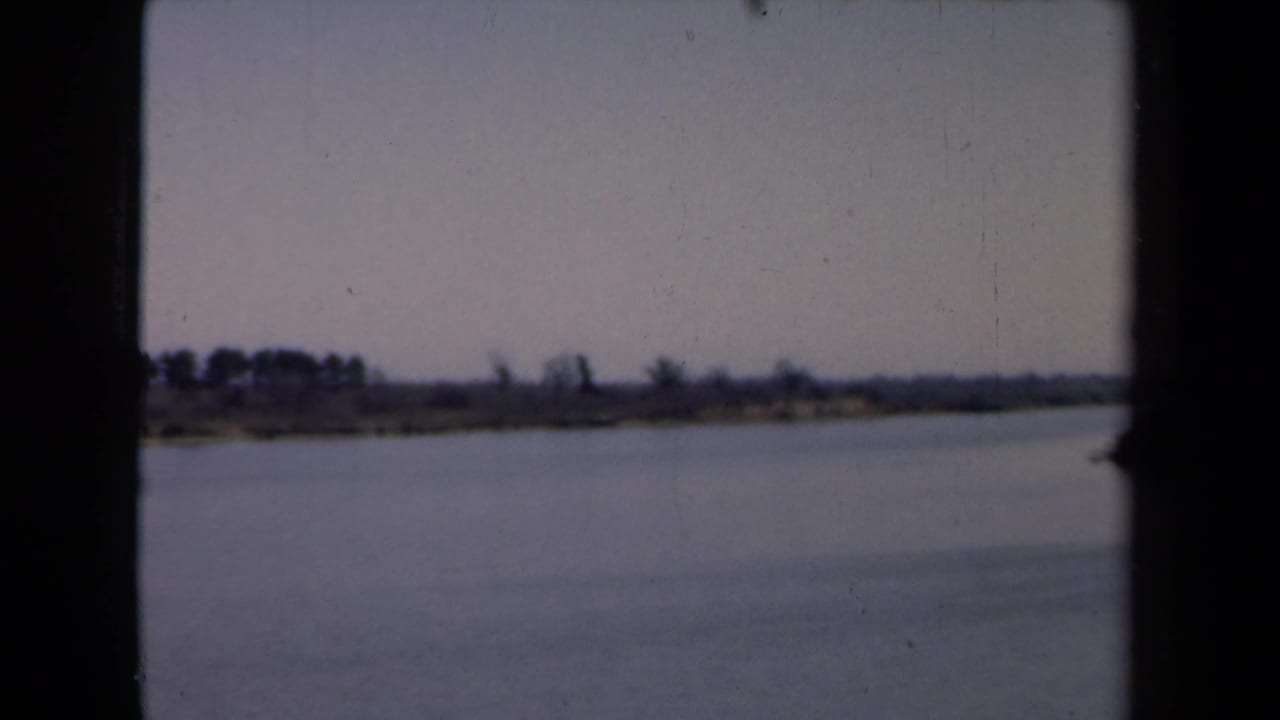 1946 Netherlands home-movie frame showing calm water, a flat distant shoreline, muted sky, and dark film gate edges.