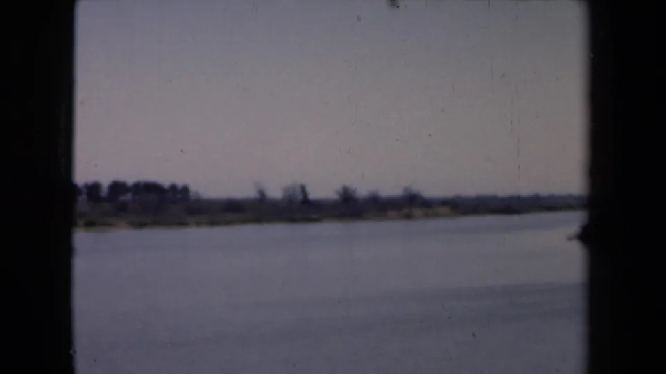 1946 Netherlands home-movie frame showing calm water, a flat distant shoreline, muted sky, and dark film gate edges.