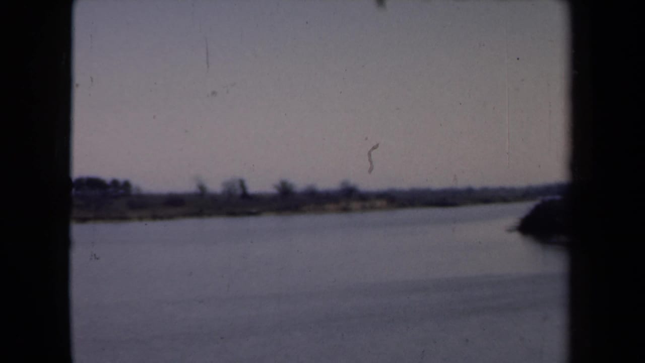Soft 8mm frame of a Dutch waterside landscape, with calm water, distant trees, pale sky, and heavy gate-edge shadows.