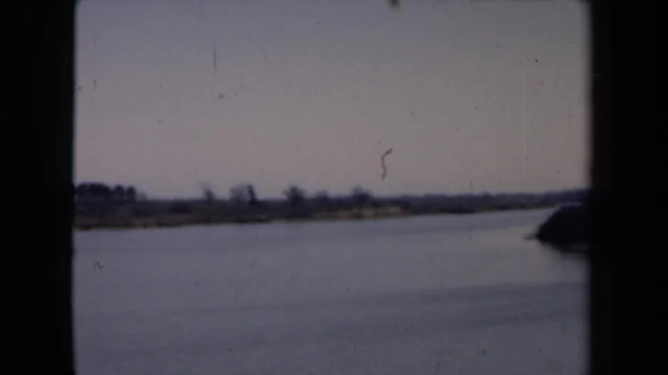 Soft 8mm frame of a Dutch waterside landscape, with calm water, distant trees, pale sky, and heavy gate-edge shadows.