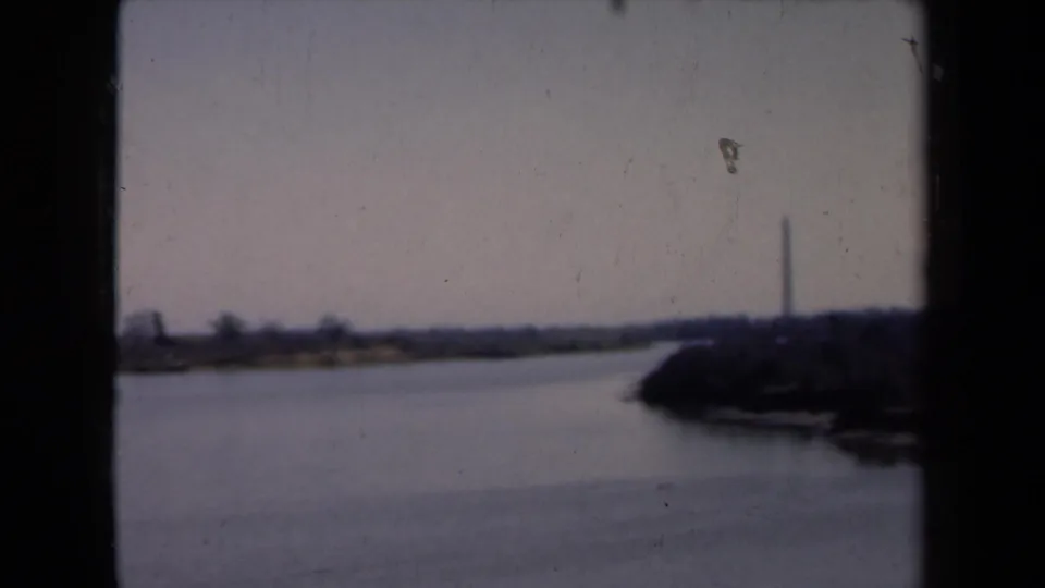 1940s 8mm frame showing a broad waterway, dark shoreline, and distant obelisk-like monument, with heavy frame edges visible.