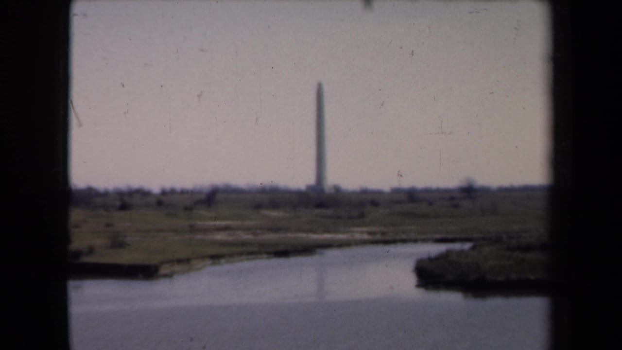A distant obelisk monument stands beyond calm water and low grassy land in visitor-shot 8mm footage.