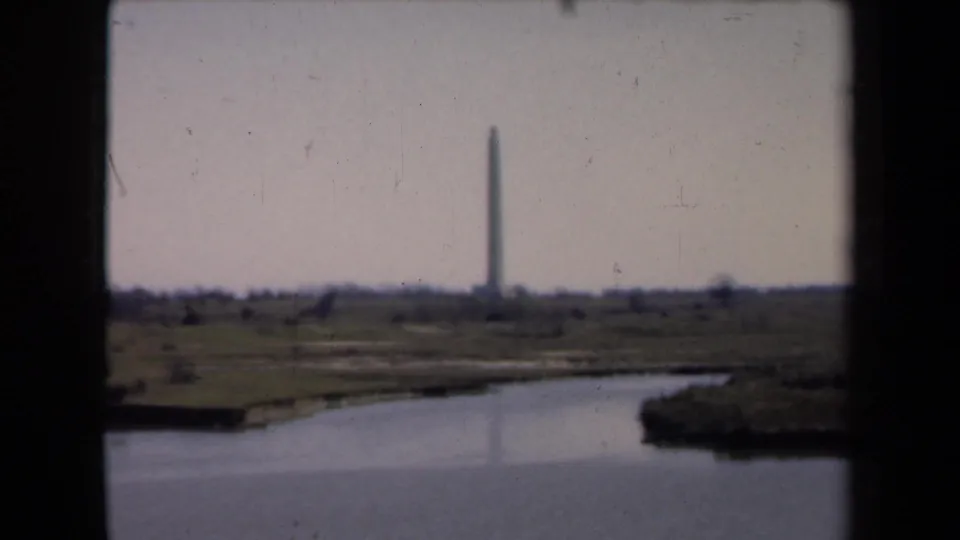 A distant obelisk monument stands beyond calm water and low grassy land in visitor-shot 8mm footage.
