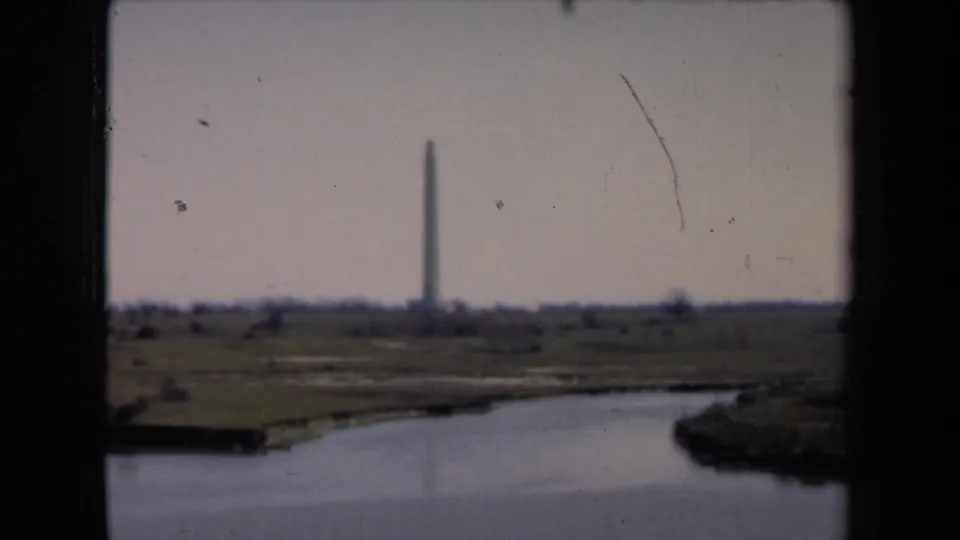 1946 8mm home-movie frame from the Netherlands metadata showing a distant tall monument across water with grassy banks and dark foreground framing.